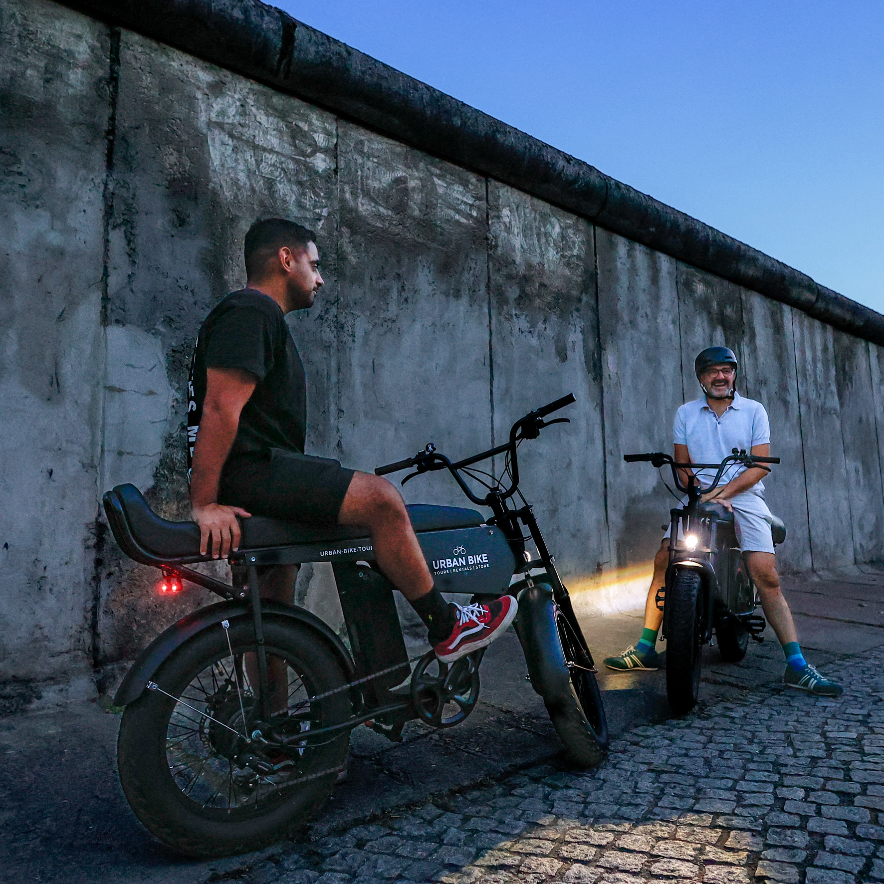 2 Personen sitzen auf ihren E-Cruisern in der Abenddämmerung vor der Berliner Mauer an der Bernauerstrasse